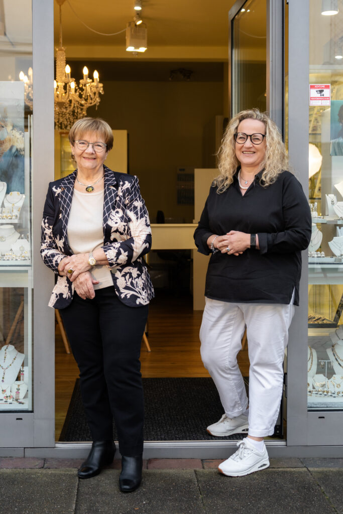 Zwei lächelnde Frauen stehen vor dem geöffneten Eingang des Schmuckgeschäfts Die SchmuckPerle, neben einer großen Schaufensterauslage voller Schmuckstücke.