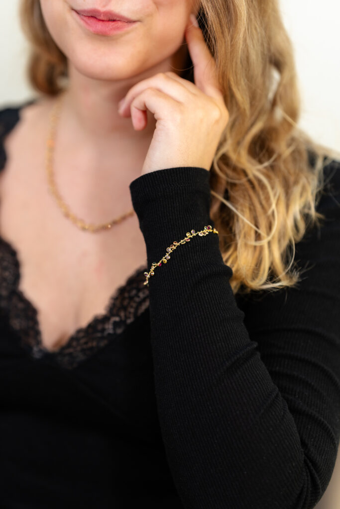 Nahaufnahme eines goldfarbenen Armbands mit kleinen farbigen Steinen, getragen über dem Ärmel eines schwarzen Oberteils; die Person legt eine Hand an ihr Gesicht und trägt zusätzlich eine passende Halskette.