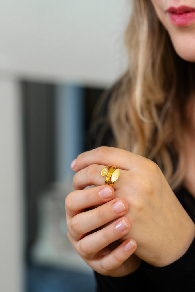 Nahaufnahme einer Frau, deren Hände locker verschränkt sind; sie trägt einen modernen goldfarbenen Ring mit einem runden Stein und einer matt strukturierten, ovalen Elementfläche.