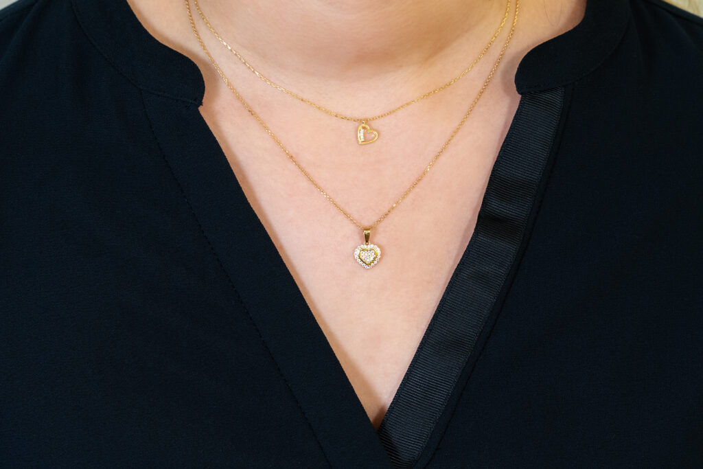 Nahaufnahme von zwei goldenen Halsketten, die übereinander getragen werden: Die obere trägt einen filigranen offenen Herzanhänger, die untere ein Herz mit funkelnden Steinen.