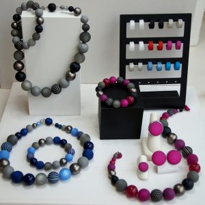 Schmuckset aus matten und glänzenden Kugelperlen in Grau, Blau, Pink und Schwarz. Zu sehen sind mehrere Halsketten, Armbänder, Ringe und Ohrstecker, teils mit gestreiften oder gemusterten Perlen.